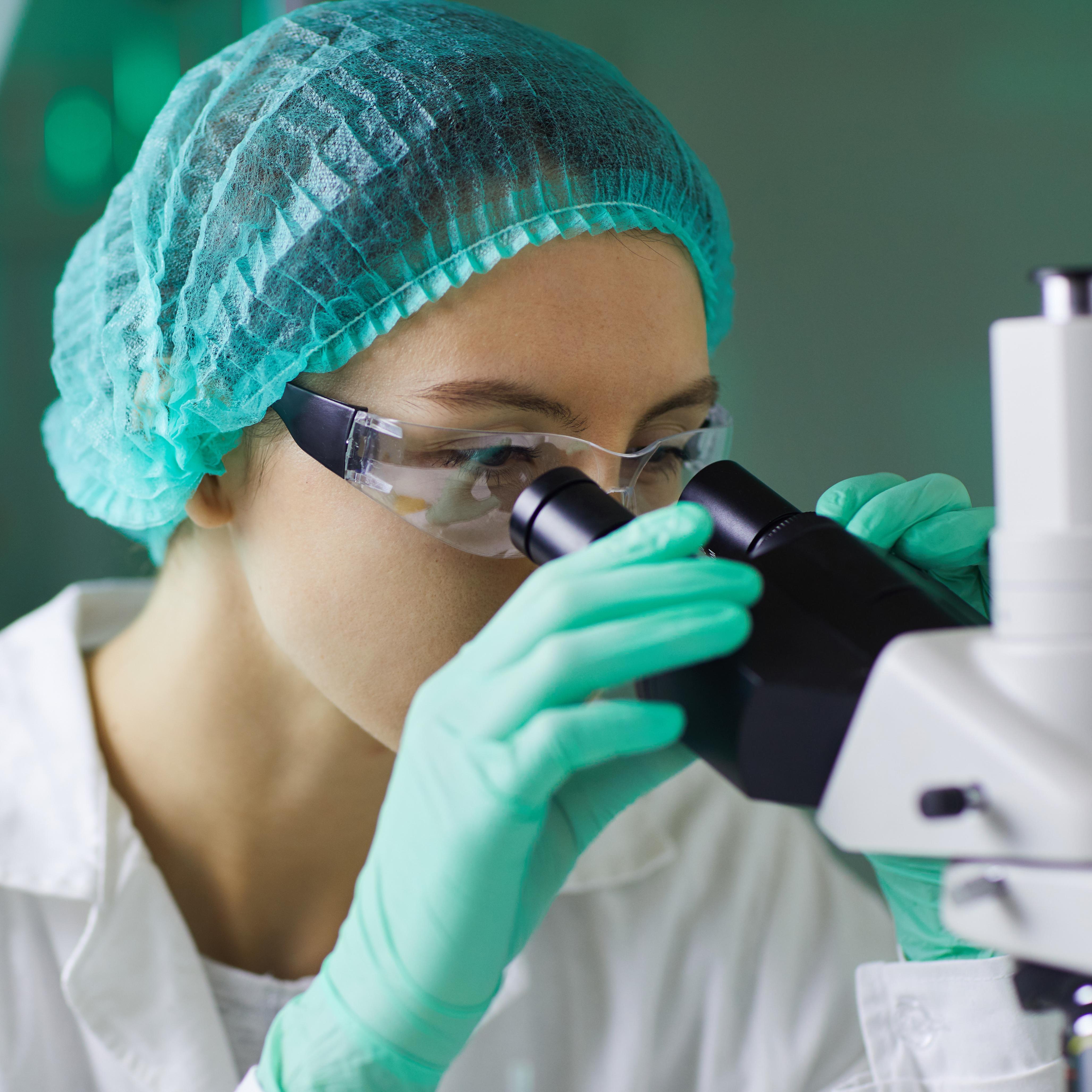 A woman adorned with personal protective equipment looks through a microscope doing medical research